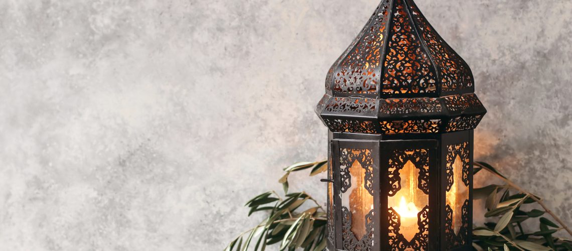Black ornamental Moroccan, Arabic lantern. Green olive leaves, branches on old wooden table, blurred grunge wall background,. Greeting card for Muslim holiday Ramadan Kareem, festive still life.